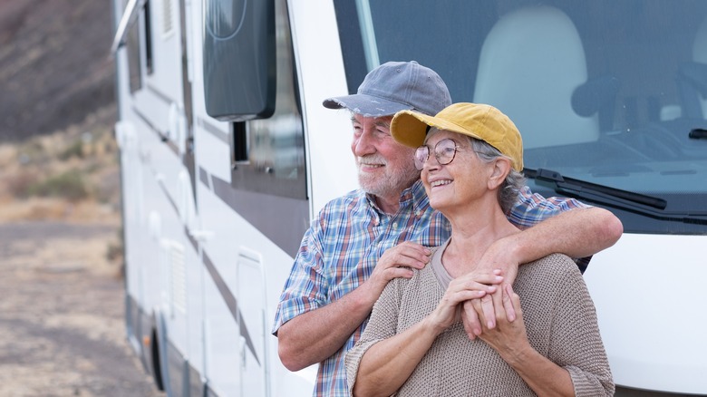 Cheerful senior couple on leisure trip standing outside a camper hugging each other and looking away.