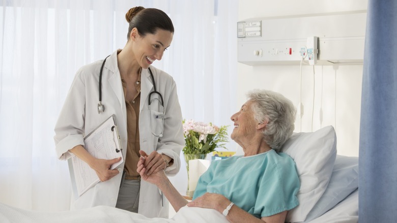 An elderly female patient in the hospital receiving medical care from a doctor holding a clipboard.