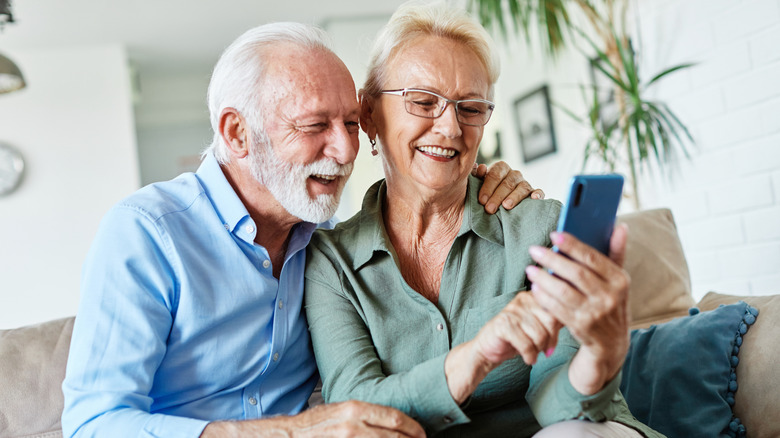 A happy retired couple sitting together on a couch looking at a phone the woman is holding and pointing to.
