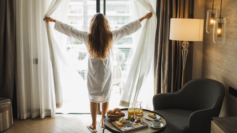 Woman opening curtains in hotel room during breakfast