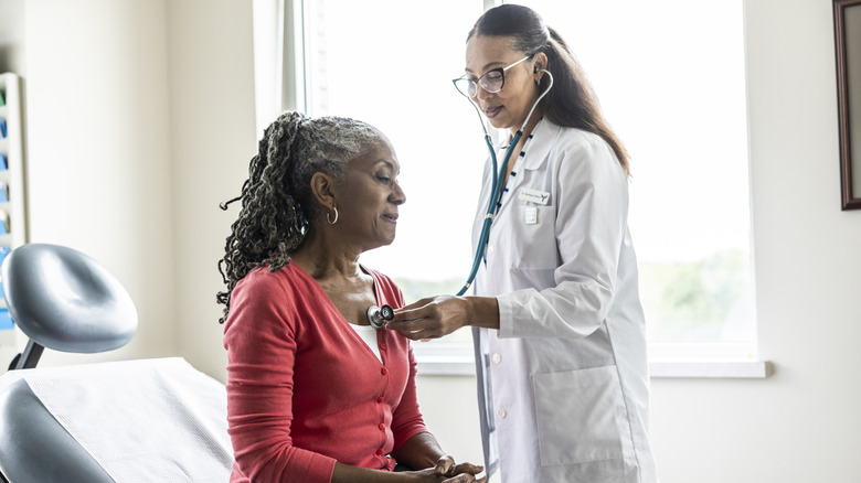 Female doctor listening to senior woman's heart in exam room