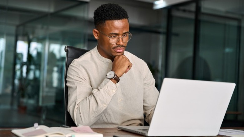 A young man with glasses sitting at a desk and funding his IRA account on a laptop.