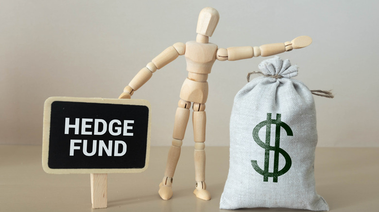 Wooden figurine holding a black sign with "hedge fund" written on it in one hand and a bag of money in the other