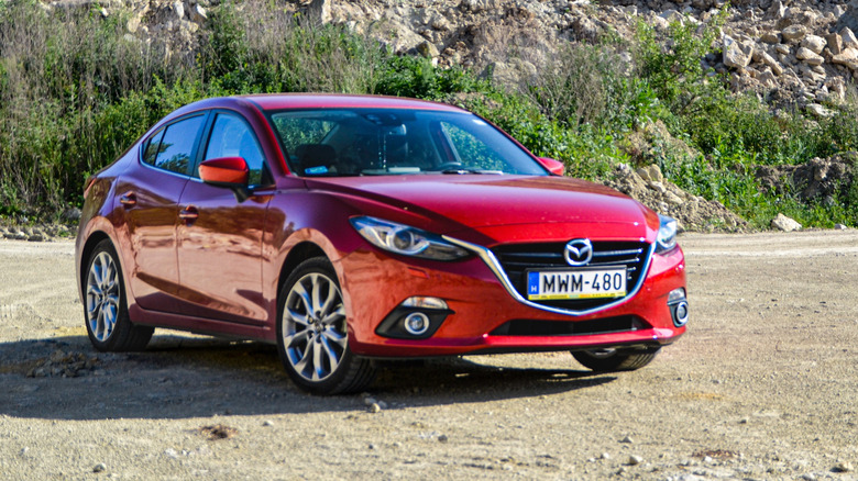 A red Mazda 3 Sedan in a sunny, rural landscape with dirt roads and gentle grassy hills.