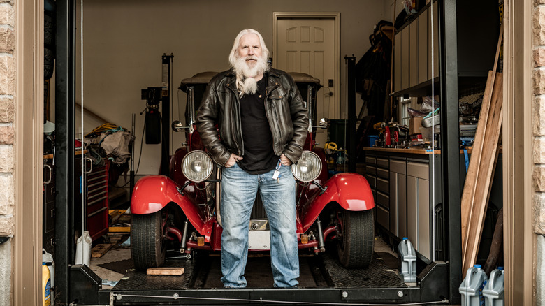 An elderly man standing in front of a vintage car in his garage.