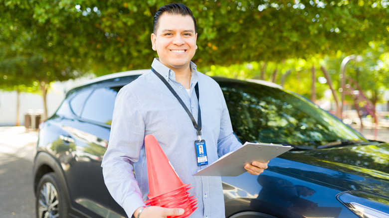 A smiling driving instructor holding traffic cones and a clipboard while standing next to a car outdoors.