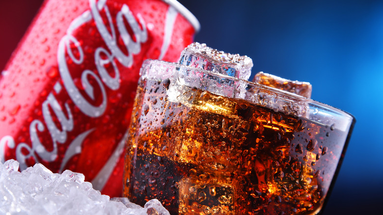 Glass of soda with ice cubes in pile of ice in front of wet Coca-Cola can