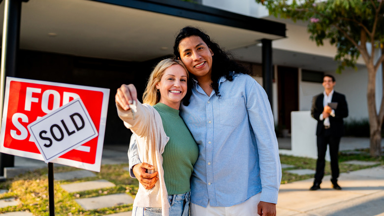 embracing couple holding a key are standing in front of Sold sign on front lawn