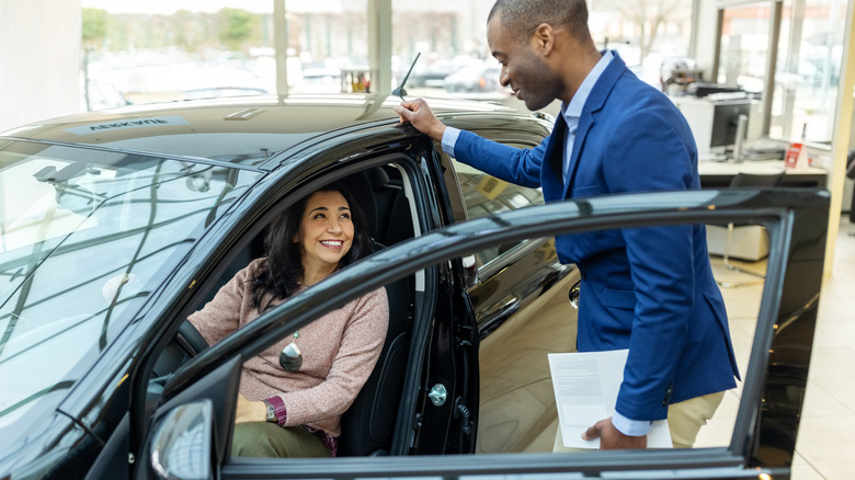 Smiling middle-aged woman sitting in display car's driver's seat talking to young car salesman