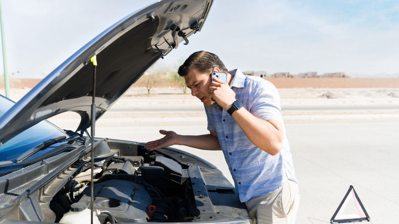 person having car trouble standing in front of the popped hood with phone to his ear