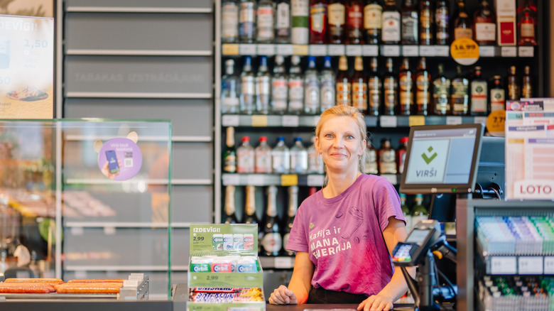 Smiling female cashier at a Virši gas station store counter with shelves of alcohol bottles in the background.