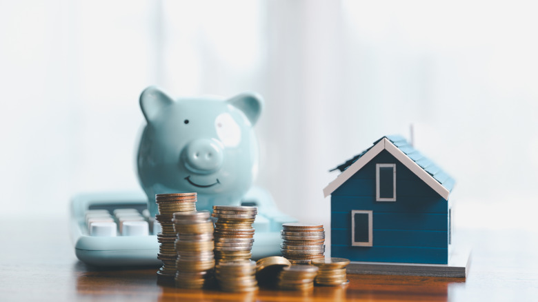 Stacks of coins, a piggy bank, a calculator, and a small model house arranged on a tabletop.