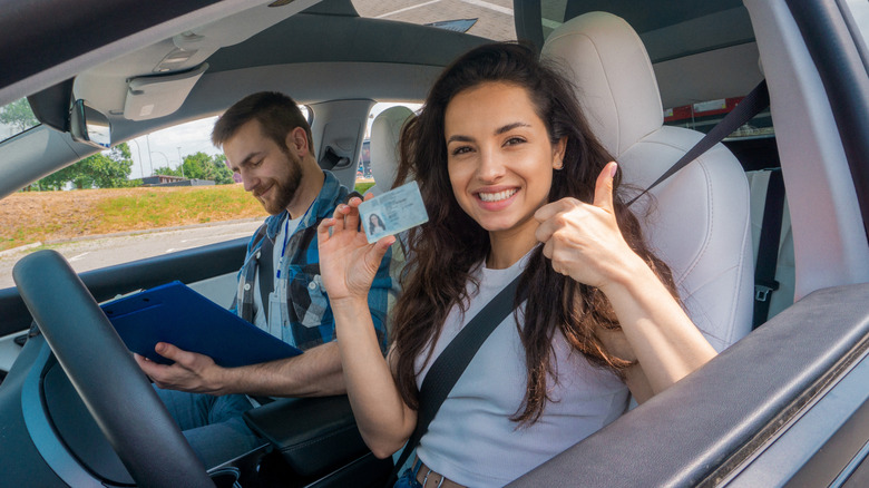 Smiling young woman in the driver seat holding her license with a driving instructor in the passenger seat