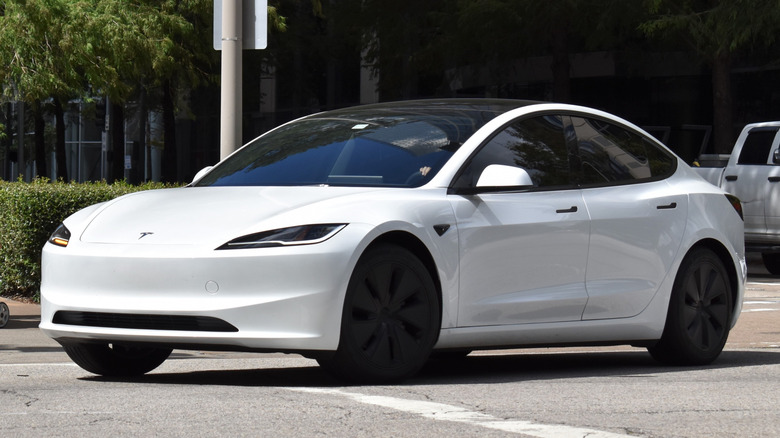 A portrait of a white Tesla Model 3 EV cruising in a downtown district.