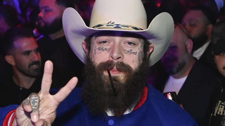 Post Malone wearing white had and smiling while making peace sign