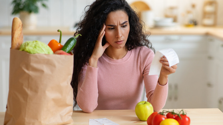 A concerned woman looks at a grocery receipt while sitting next to a full paper grocery bag at a table in a bright kitchen