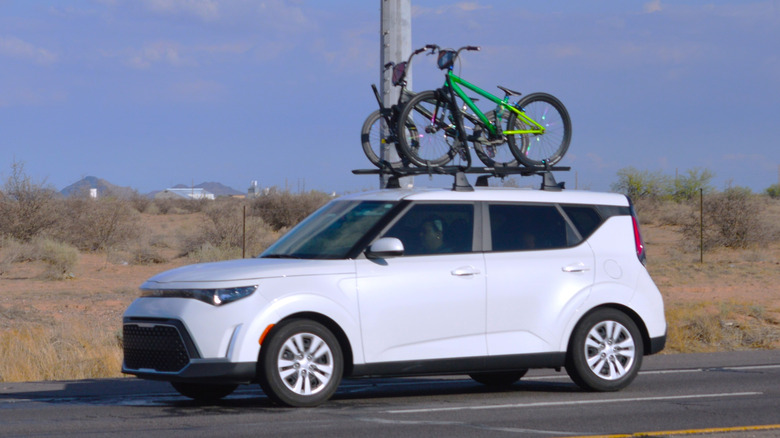 White Kia Soul driving along a rural desert highway with roof‑mounted bicycles in the Sonoran Desert.