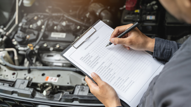 A mechanic with a checklist on a clipboard standing over a car with its hood open.