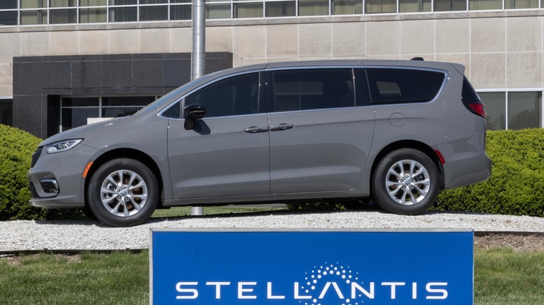 Gray Chrysler minivan parked on a display area outside an office building, with a blue Stellantis sign in the foreground