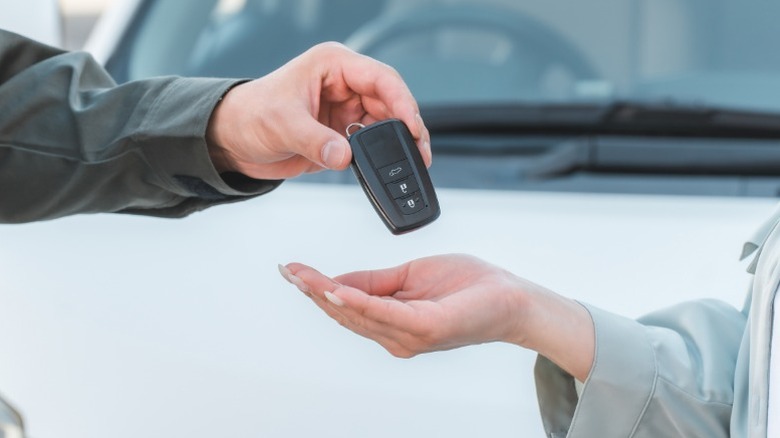 Hands showing a person handing over car keys to a buyer in front of a white vehicle.