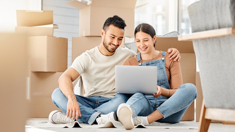 A couple with moving boxes around them looking at a laptop.