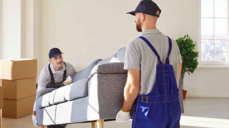 Two men in overalls delivering a new couch by bringing it into a living room with moving boxes visible.
