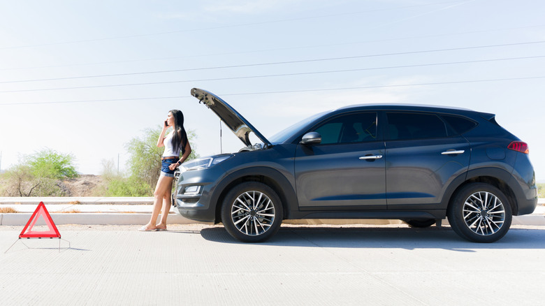 young woman on phone in front of blue SUV with raised hood