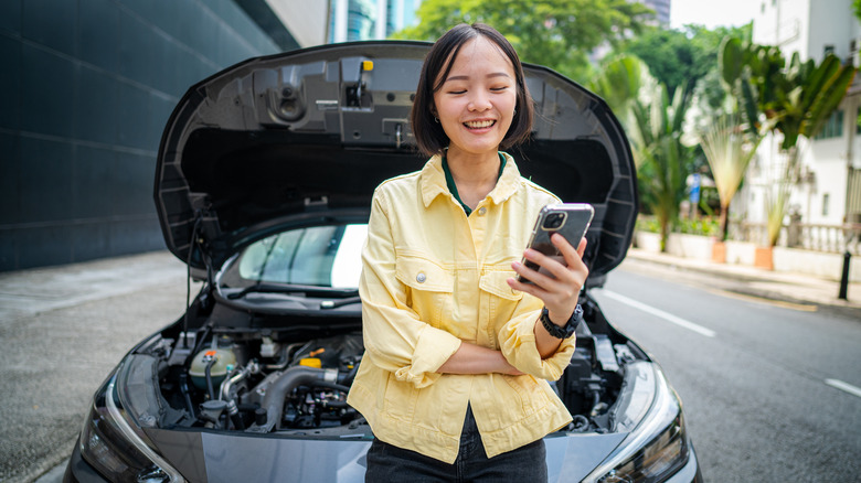 A smiling woman waiting for assistance near her stalled vehicle on the roadside, after a response from the insurance company.