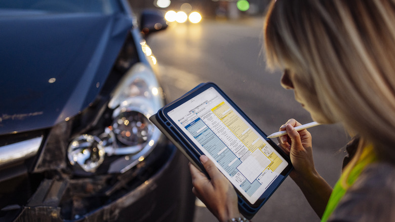 woman wearing a reflective safety vest writes on a digital tablet near a damaged car after a traffic accident.
