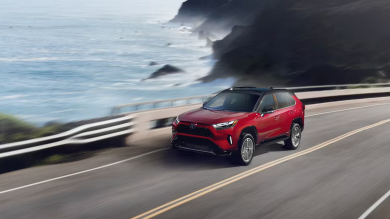 a red 2025 Toyota RAV4 Plug-in Hybrid travels the curve in a highway hugging a rocky seacoast