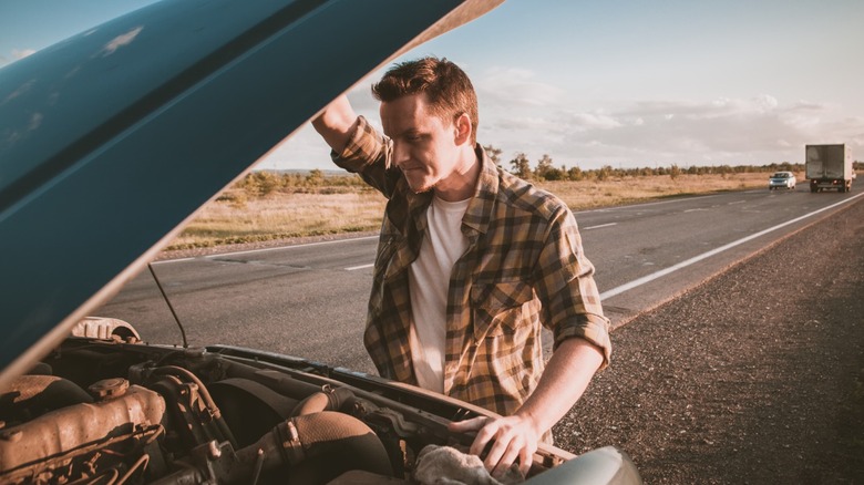 a man looks at his car's engine beside a highway