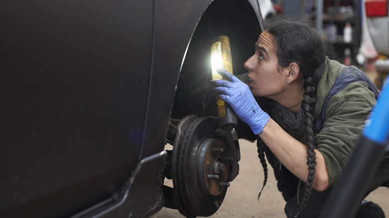 A female mechanic examines the tire-area of a car with a light.