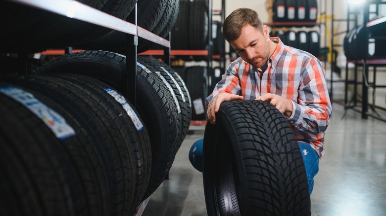Man inspecting a tire tread while kneeling in a tire shop aisle lined with stacked tires