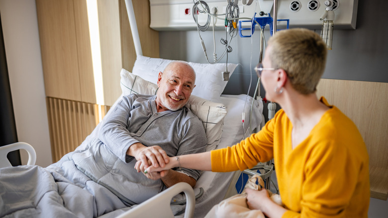 A smiling man laying in a hospital bed holding hands with a visitor in the hospital.
