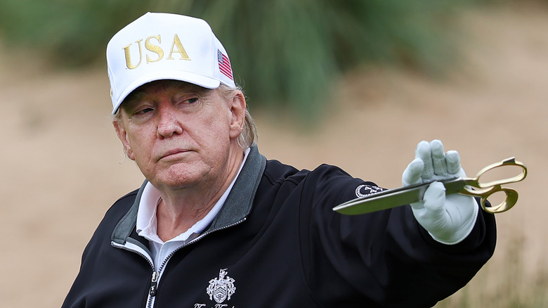 President Donald Trump wearing white USA cap and  Trump Turnberry outfit while holding a pair of golden scissors.