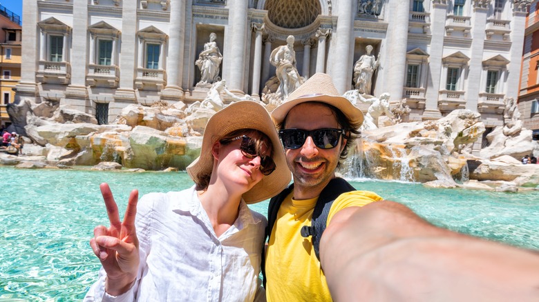 A millennial couple smiling while taking a selfie in fron of the Trevi Fountain in Rome, Italy.