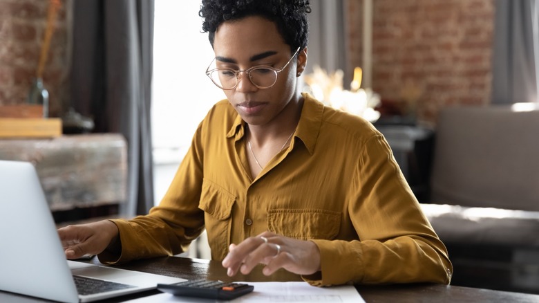 A young woman typing on a calculator while sitting in front of a laptop doing work.