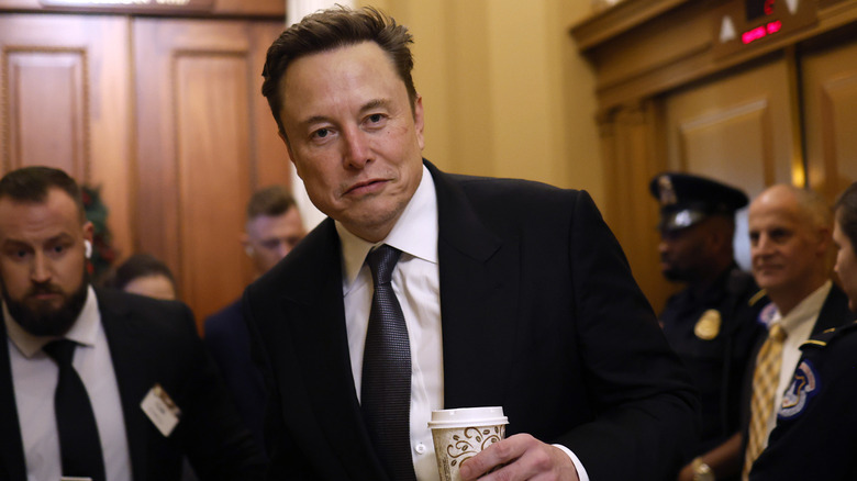 Elon Musk holds a cup of coffee in a wood-paneled corridor, looking tired.