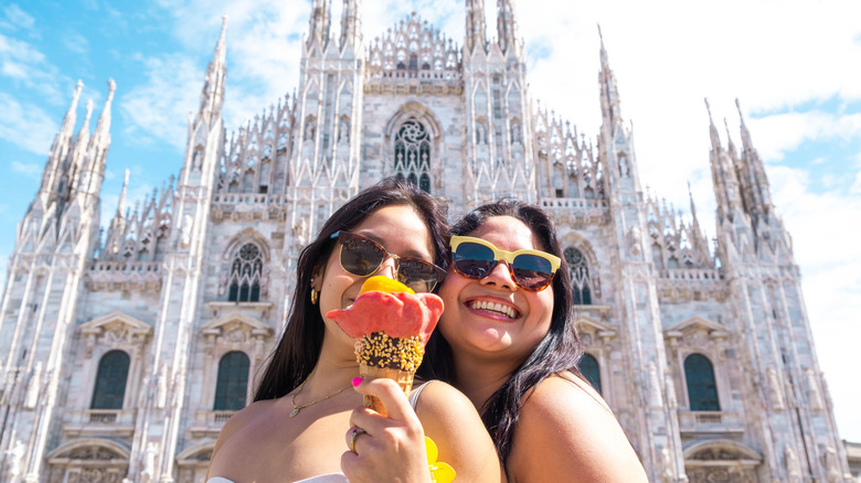 Two women tasting a creative handmade gelato with rose shape and colors in front of the Duomo of Milan, Italy.