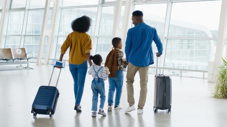 A family with two adults and two children walking in an airport terminal waiting area with two suitcases.