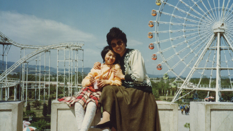 A mother and daughter smiling together at an amusement park with a Ferris wheel and roller coaster in the background