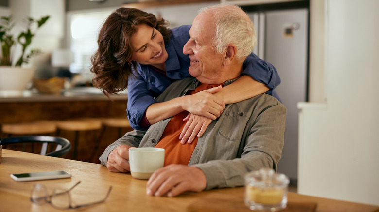 an adult daughter hugs her seated gray-haired father from behind