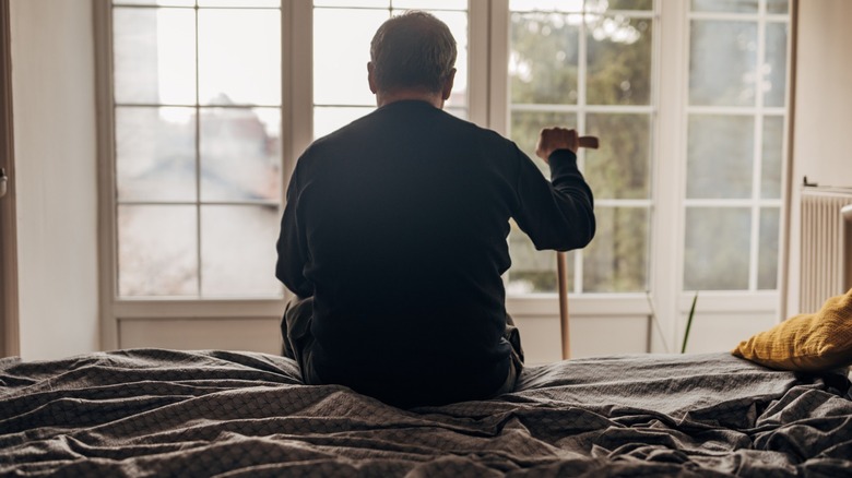 An older man with a cane sitting on a bed in a retirement home.