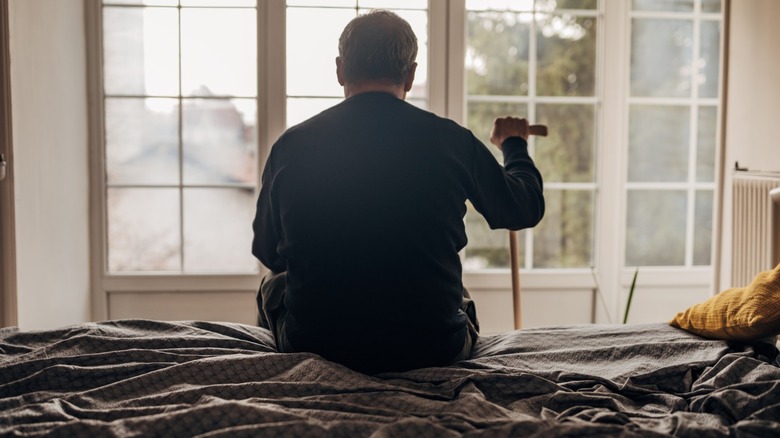 An older man with a cane sitting on a bed in a retirement home.