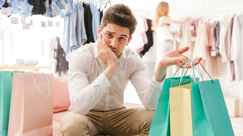 An unhappy man sitting in a clothing store holding multiple shopping bags.