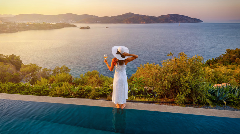 A woman stands on the edge of a pool watching the sun set over the ocean
