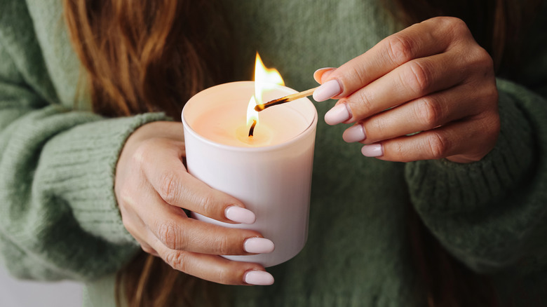 Woman wearing long sleeve shirt with polished nails holds and lights a thick candle
