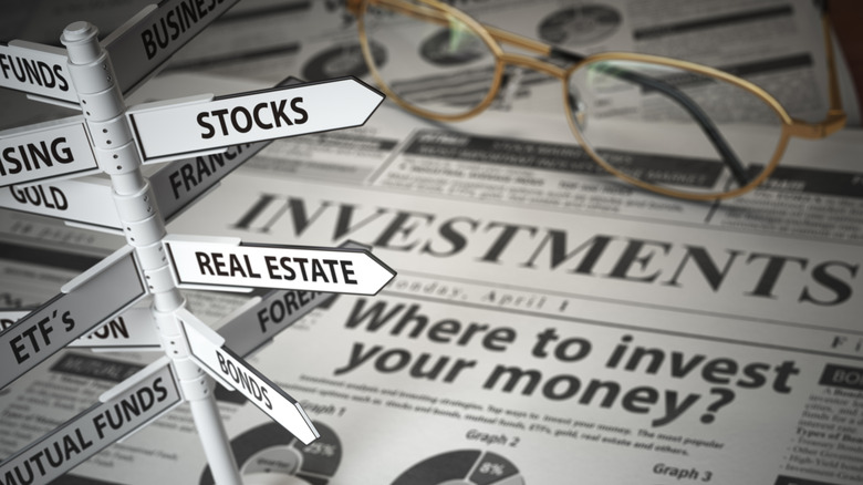 A signpost displaying different investment tools on top of a newspaper with glasses in the background