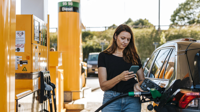 Slighting frowning woman pumping gas and looking at her phone