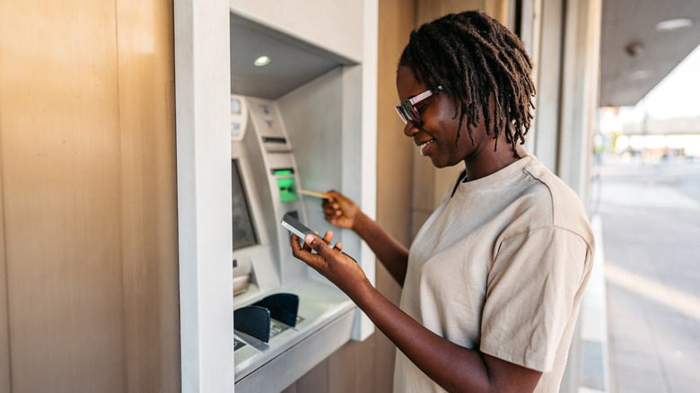 A young woman standing outside of an ATM as she checks her phone